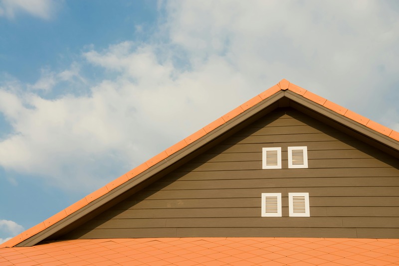 Close-up of a gable roof with orange tiles and two pairs of white vent windows against a blue sky with light clouds